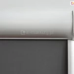 Zbliżenie na górny profil rolety wewnętrznej w białej, zaokrąglonej kasecie osłaniającej mechanizm nawojowy. Pod kasetą znajduje się gładka tkanina w kolorze ciemnoszarym lub antracytowym o gęstym, jednolitym splocie, który zapewnia maksymalne zaciemnienie pomieszczenia. Po prawej stronie widoczny jest biały łańcuszek koralikowy służący do sterowania wysokością rolety, który wychodzi z bocznego elementu wykończeniowego kasety. Pionowa, biała prowadnica boczna biegnie w dół wzdłuż krawędzi materiału, zapewniając jego stabilne przyleganie. Całość konstrukcji, obejmująca kasetę, boczki oraz prowadnice, jest wykonana z białego tworzywa lub lakierowanego aluminium o satynowym wykończeniu. Tło zdjęcia jest jasne i neutralne, co uwydatnia techniczne detale montażowe oraz kontrast między jasną obudową a ciemnym materiałem przesłonowym.