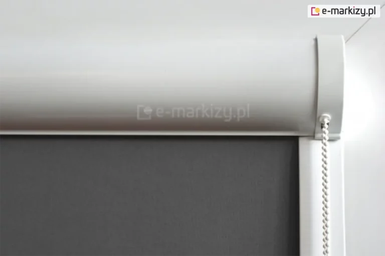 Zbliżenie na górny profil rolety wewnętrznej w białej, zaokrąglonej kasecie osłaniającej mechanizm nawojowy. Pod kasetą znajduje się gładka tkanina w kolorze ciemnoszarym lub antracytowym o gęstym, jednolitym splocie, który zapewnia maksymalne zaciemnienie pomieszczenia. Po prawej stronie widoczny jest biały łańcuszek koralikowy służący do sterowania wysokością rolety, który wychodzi z bocznego elementu wykończeniowego kasety. Pionowa, biała prowadnica boczna biegnie w dół wzdłuż krawędzi materiału, zapewniając jego stabilne przyleganie. Całość konstrukcji, obejmująca kasetę, boczki oraz prowadnice, jest wykonana z białego tworzywa lub lakierowanego aluminium o satynowym wykończeniu. Tło zdjęcia jest jasne i neutralne, co uwydatnia techniczne detale montażowe oraz kontrast między jasną obudową a ciemnym materiałem przesłonowym.