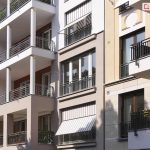 Zdjęcie przedstawia fragment wielokondygnacyjnego budynku mieszkalnego o zróżnicowanej architekturze elewacji, na której zainstalowano markizorolety zewnętrzne. W centralnej części kadru widoczny jest pionowy pas jasnobeżowej fasady z dwoma dużymi oknami balkonowymi, wyposażonymi w osłony z tkaniny w klasyczne, pionowe biało-szare pasy. Dolna markizoroleta jest rozwinięta, a jej końcowa część została odchylona pod kątem na zewnątrz za pomocą bocznych ramion, tworząc daszek nad czarną, metalową balustradą. Górna osłona w tym samym pasie pozostaje niemal całkowicie zwinięta w kasecie. Po lewej stronie widoczny jest segment budynku z głębokimi loggiami wspartymi na białych kolumnach ze złotymi detalami, natomiast po prawej stronie znajduje się fragment elewacji o barwie piaskowej z ozdobnymi gzymsami i klasycznymi balkonami. Całość ukazana jest w świetle dziennym, a w lewym górnym rogu widoczne są liście drzewa rzucające delikatny cień na budynek.