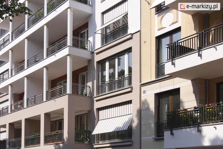 Zdjęcie przedstawia fragment wielokondygnacyjnego budynku mieszkalnego o zróżnicowanej architekturze elewacji, na której zainstalowano markizorolety zewnętrzne. W centralnej części kadru widoczny jest pionowy pas jasnobeżowej fasady z dwoma dużymi oknami balkonowymi, wyposażonymi w osłony z tkaniny w klasyczne, pionowe biało-szare pasy. Dolna markizoroleta jest rozwinięta, a jej końcowa część została odchylona pod kątem na zewnątrz za pomocą bocznych ramion, tworząc daszek nad czarną, metalową balustradą. Górna osłona w tym samym pasie pozostaje niemal całkowicie zwinięta w kasecie. Po lewej stronie widoczny jest segment budynku z głębokimi loggiami wspartymi na białych kolumnach ze złotymi detalami, natomiast po prawej stronie znajduje się fragment elewacji o barwie piaskowej z ozdobnymi gzymsami i klasycznymi balkonami. Całość ukazana jest w świetle dziennym, a w lewym górnym rogu widoczne są liście drzewa rzucające delikatny cień na budynek.