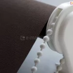 Zbliżenie uchwytu rolety Plus w białym kolorze z mechanizmem łańcuszkowym do zwijania i rozwijania rolety z brązową tkaniną. Roleta plus dzień noc posiada odmienny system mocowania i sterowania.