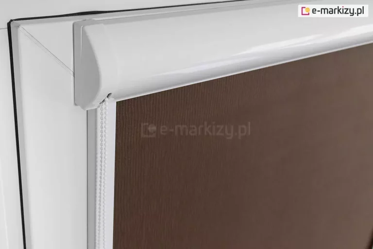 Lewy bok kasety przestrzennej w kolorze białym zamocowanej na skrzydle okna o białej ramie. Z kasety wystaje łańcuszek do sterowania roletą. Widoczna także przyciemniająca tkanina rolety w kolorze brązowym.