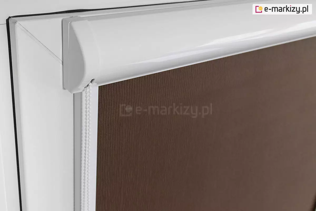Lewy bok kasety przestrzennej w kolorze białym zamocowanej na skrzydle okna o białej ramie. Z kasety wystaje łańcuszek do sterowania roletą. Widoczna także przyciemniająca tkanina rolety w kolorze brązowym.