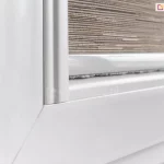 Zbliżenie na dolny narożnik białej ramy okiennej PCV z zamontowaną na oknie roletką tkaninową. Zdjęcie ukazuje detal zakończenia osłony, gdzie pionowa, biała prowadnica boczna styka się z poziomą listwą dolną materiału. Tkanina rolety charakteryzuje się gęstym, poziomym splotem w odcieniach beżu i brązu, przypominającym strukturę naturalnej maty lub lnu. Dolna listwa obciążająca tkaninę jest wyposażona w dyskretną uszczelkę szczotkową, która przylega do ramy okna, minimalizując prześwity i chroniąc przed kurzem. Całość systemu jest precyzyjnie dopasowana do profilu skrzydła okiennego, co gwarantuje stabilne prowadzenie materiału podczas podnoszenia i opuszczania.