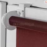 Roleta mini z uchwytem zawieszanym na skrzydle okna o białym kolorze. Widoczna także tkanina rolety w kolorze czerwono-brązowym.