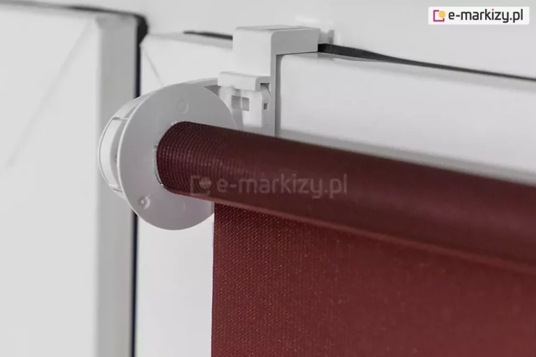 Roleta mini z uchwytem zawieszanym na skrzydle okna o białym kolorze. Widoczna także tkanina rolety w kolorze czerwono-brązowym.