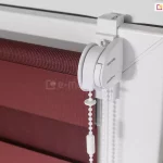 Zbliżenie narożnika okna z zawiasem o białej ramie, na której zawieszona została mocowaniem bezinwazyjnym roletka mini z tkaniną dzień noc w pasy w kolorze czerwonego wina.