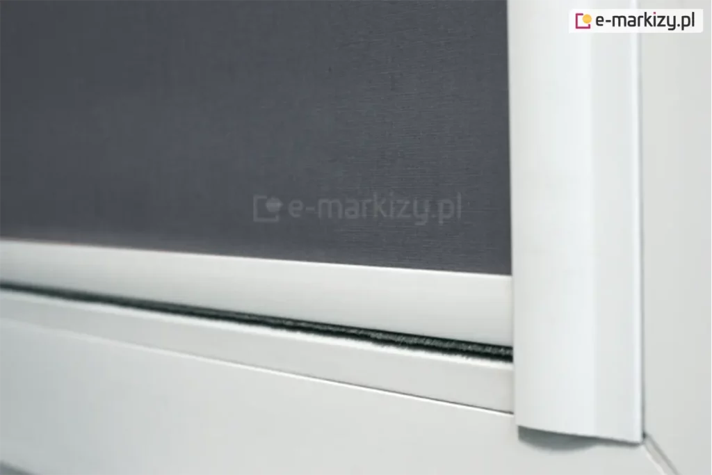 Zbliżenie prowadnicy płaskiej rolety materiałowej LUX z widocznym dolnym obciążnikiem z uszczelką szczotkową. Biała konstrukcja rolety jest najczęściej wybieranym rozwiązaniem dla rolet, ponieważ dopasowuje się do białym ram okiennych. W rozwiniętej rolecie zastosowano szarą tkaninę zaciemniającą, która wraz z prowadnicami przyklejonymi do listew przyszybowych skutecznie przyciemnia pomieszczenie.