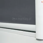 Zbliżenie prowadnicy płaskiej rolety materiałowej LUX z widocznym dolnym obciążnikiem z uszczelką szczotkową. Biała konstrukcja rolety jest najczęściej wybieranym rozwiązaniem dla rolet, ponieważ dopasowuje się do białym ram okiennych. W rozwiniętej rolecie zastosowano szarą tkaninę zaciemniającą, która wraz z prowadnicami przyklejonymi do listew przyszybowych skutecznie przyciemnia pomieszczenie.