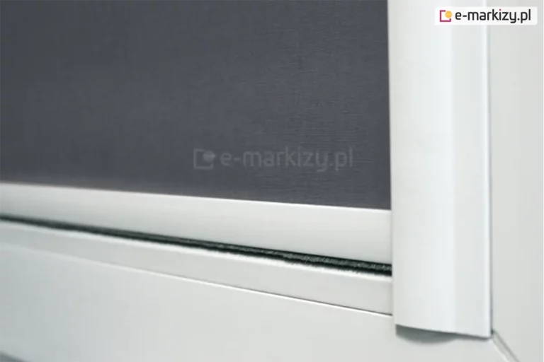 Zbliżenie prowadnicy płaskiej rolety materiałowej LUX z widocznym dolnym obciążnikiem z uszczelką szczotkową. Biała konstrukcja rolety jest najczęściej wybieranym rozwiązaniem dla rolet, ponieważ dopasowuje się do białym ram okiennych. W rozwiniętej rolecie zastosowano szarą tkaninę zaciemniającą, która wraz z prowadnicami przyklejonymi do listew przyszybowych skutecznie przyciemnia pomieszczenie.