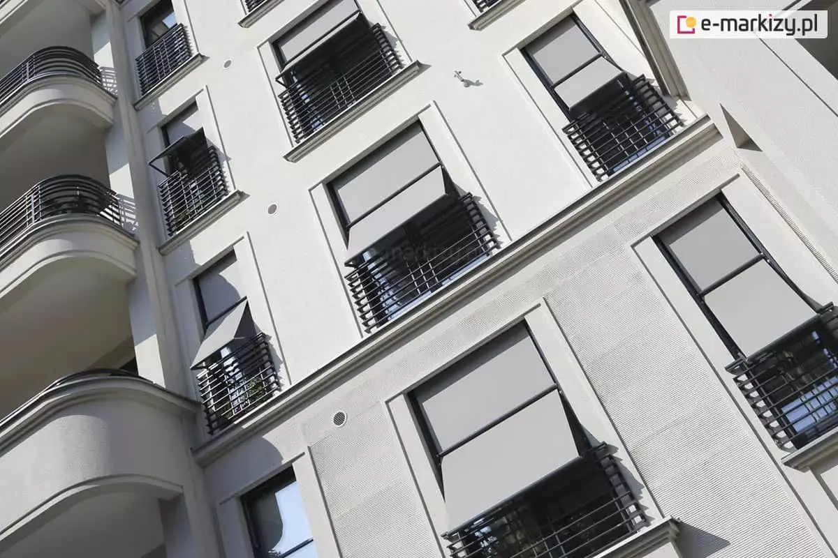 Zdjęcie prezentuje perspektywę z dołu na jasną, tynkowaną elewację nowoczesnego budynku wielorodzinnego, na której zamontowano liczne markizorolety zewnętrzne w kolorze szarym. Osłony są zainstalowane w pionowych rzędach okien i znajdują się w różnym stopniu rozwinięcia. Większość markizorolet jest częściowo opuszczona, a ich dolna część została odchylona na zewnątrz za pomocą bocznych ramion, co tworzy funkcjonalny daszek nad oknami i balkonami. Systemy te są zintegrowane z czarnymi, ażurowymi balustradami zabezpieczającymi portfenetry. Po lewej stronie kadru widoczne są zaokrąglone balkony z pasującymi, ciemnymi barierkami o poziomym układzie prętów. Elewacja posiada subtelne boniowania i pionowe podziały, które nadają strukturę fasadzie. Całość jest oświetlona silnym światłem dziennym, podkreślającym techniczne detale konstrukcji osłon oraz kontrast między szarym materiałem a białą ścianą budynku.