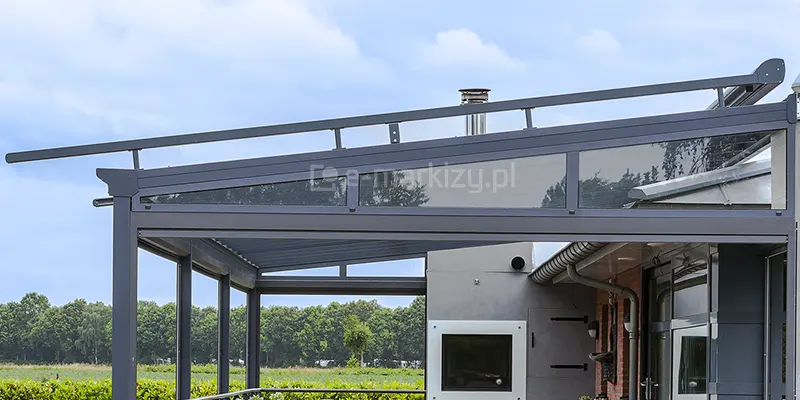 Szklane zadaszenie tarasu model Terano wyposażone w zewnętrzny system ochrony przeciwsłonecznej w postaci rolety poziomej montowanej nad konstrukcją. Całość oparta jest na profilach aluminiowych w kolorze antracytowym. Nad szklanym dachem zadaszenia widoczna jest metalowa podbudowa z prowadnicami, na których opiera się mechanizm rolety, co pozwala na stworzenie dystansu zapewniającego cyrkulację powietrza między szkłem a tkaniną. Boczna ściana zadaszenia została wypełniona stałym przeszkleniem o trójkątnym kształcie (tzw. klinem), który domyka przestrzeń pod skosem dachu. Poniżej klina zamontowano poziomą kasetę rolety bocznej lub screena. Konstrukcja jest zintegrowana z bryłą budynku o mieszanej elewacji (cegła i szary tynk), w której widoczny jest komin oraz system rynnowy. W tle rozpościera się widok na otwartą przestrzeń zieloną z linią drzew, co podkreśla funkcjonalność osłon przeciwsłonecznych w mocno nasłonecznionym terenie.