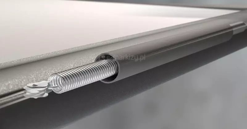 Grafika przedstawia szczegółowy przekrój technologiczny elementu napinającego tkaninę w systemie osłony przeciwsłonecznej typu Veranda. Centralnym punktem obrazu jest metalowa, spiralna sprężyna naciągowa, która jest częściowo wsunięta wewnątrz profilu aluminiowego. Sprężyna ta pełni kluczową rolę w mechanizmie naciągu tkaniny, zapewniając jej odpowiednie napięcie podczas rozwijania i zwijania osłony, co przekłada się na płynną pracę systemu i estetyczny wygląd tkaniny bez marszczeń. Prezentacja ta w sposób poglądowy ilustruje dbałość o detale konstrukcyjne oraz zaawansowanie techniczne mechanizmu ukrytego w profilach, które odpowiadają za stabilność i trwałość całej osłony.