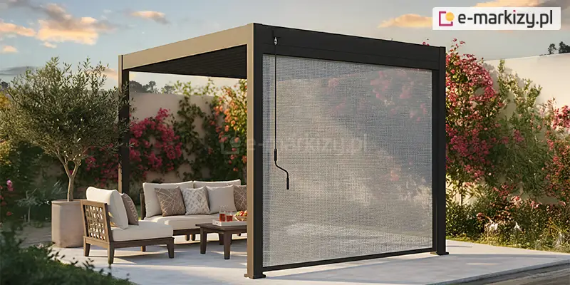 Grafika przedstawia wizualizację nowoczesnej pergoli ogrodowej w aranżacji tarasowej, uchwyconą w ciepłym świetle zachodzącego słońca. Konstrukcja pergoli jest wolnostojąca, wykonana z profili aluminiowych w głębokim, matowym kolorze antracytowym. Głównym elementem prezentowanym na zdjęciu jest pionowa roleta boczna typu screen, zamontowana na froncie konstrukcji. Tkanina rolety ma jasnoszary kolor i strukturę drobnej siatki, która zapewnia przezierność od wewnątrz przy jednoczesnym ograniczaniu nagrzewania i zapewnianiu prywatności. Po lewej stronie przedniego słupa zamocowana jest długa, czarna korba służąca do ręcznego opuszczania i podnoszenia rolety. Pod zadaszeniem pergoli znajduje się strefa wypoczynkowa z wygodnymi meblami ogrodowymi, na którą składają się jasnobeżowe sofy z licznymi poduszkami dekoracyjnymi w geometryczne wzory oraz niski, drewniany stolik kawowy, na którym ustawiono szklanki z napojami. Podłoże tarasu wyłożone jest gładkimi, jasnymi płytami wielkoformatowymi. Tło aranżacji tworzy biały mur ogrodzeniowy gęsto porośnięty pnącymi roślinami o intensywnie różowych kwiatach oraz zielone drzewko oliwne w dużej, jasnej donicy po lewej stronie. Niebo w tle ma odcienie błękitu i pomarańczu, co podkreśla relaksacyjny charakter prezentowanej przestrzeni.