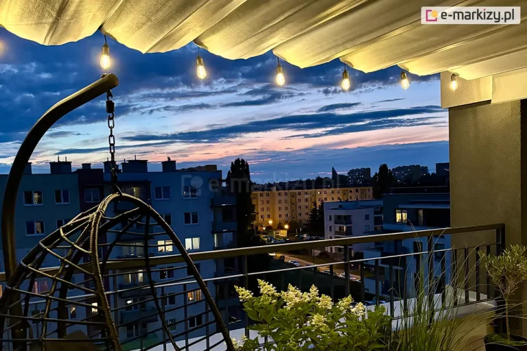 Zdjęcie przedstawia nowoczesną aranżację balkonu w bloku mieszkalnym podczas zmierzchu, gdzie głównym elementem osłonowym jest jasna pozioma roleta rzymska Supro zamontowana pod zadaszeniem. Tkanina rolety jest ułożona w charakterystyczne, regularne fale, a pod nią zawieszono girlandę świetlną z żarówkami typu retro, które emitują ciepłe, żółte światło oświetlające materiał od spodu. Po lewej stronie pierwszego planu widoczny jest fragment czarnego, wiszącego fotela ogrodowego o ażurowej konstrukcji wykonanej z technorattanu, podwieszonego na metalowym stelażu. Balustrada balkonu jest ciemna, metalowa, o prostym pionowym szczeblowaniu. Za balustradą znajdują się donice z roślinnością, w tym kwitnące na biało hortensje oraz ozdobne trawy. W tle rozpościera się panorama osiedla z wielopiętrowymi budynkami mieszkalnymi, w których widoczne są zapalone światła w oknach. Niebo nad miastem ma głęboki granatowy kolor z pasmami różu i pomarańczu pozostałymi po zachodzie słońca.