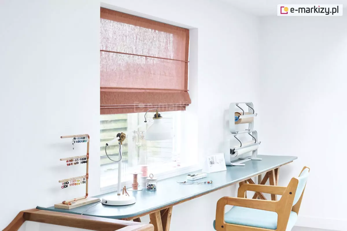 Zdjęcie przedstawia jasne i uporządkowane wnętrze pracowni lub pokoju hobby, w którym centralnym punktem jest okno przesłonięte roletą rzymską w kolorze terakotowym lub zgaszonego pomarańczu. Roleta wykonana jest z półtransparentnej tkaniny o wyraźnym, pionowym splotu, który przypomina naturalne włókna. Jest ona podciągnięta do około 1/3 wysokości okna, a jej materiał jest podzielony poziomymi fiszbinami, tworząc estetyczne zakładki w dolnej części. Przez tkaninę oraz odsłoniętą dolną część szyby wpada jasne, dzienne światło. Pod oknem znajduje się długi blat roboczy w kolorze błękitnym, wsparty na drewnianych kozłach. Na blacie, bezpośrednio przy oknie, stoi biała lampka biurkowa oraz małe akcesoria rzemieślnicze: szpulki nici i szklany słoiczek z drobnymi elementami. Po lewej stronie blatu umieszczony jest stojak na biżuterię wykonany z miedzianych rurek, na którym zawieszone są kolorowe kolczyki. Po prawej stronie blatu stoi metalowy podajnik z dwiema rolkami papieru pakowego. Przy biurku stoi drewniane krzesło z jasnobłękitnym obiciem na siedzisku i oparciu. Ściany pomieszczenia są białe i gładkie, co podkreśla czystość i nowoczesny charakter aranżacji, a kolorowa roleta stanowi główny akcent barwny we wnętrzu.