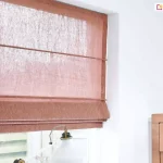Zamocowana we wnęce okiennej pomieszczenia o białych ścianach roleta rzymska model 9, z tkaniną półprzezierną w kolorze terakotowym. Przed oknem widoczna lampka biurkowa z białym kloszem. Po prawej przy ścianie widoczny narożnik drewnianej komody.