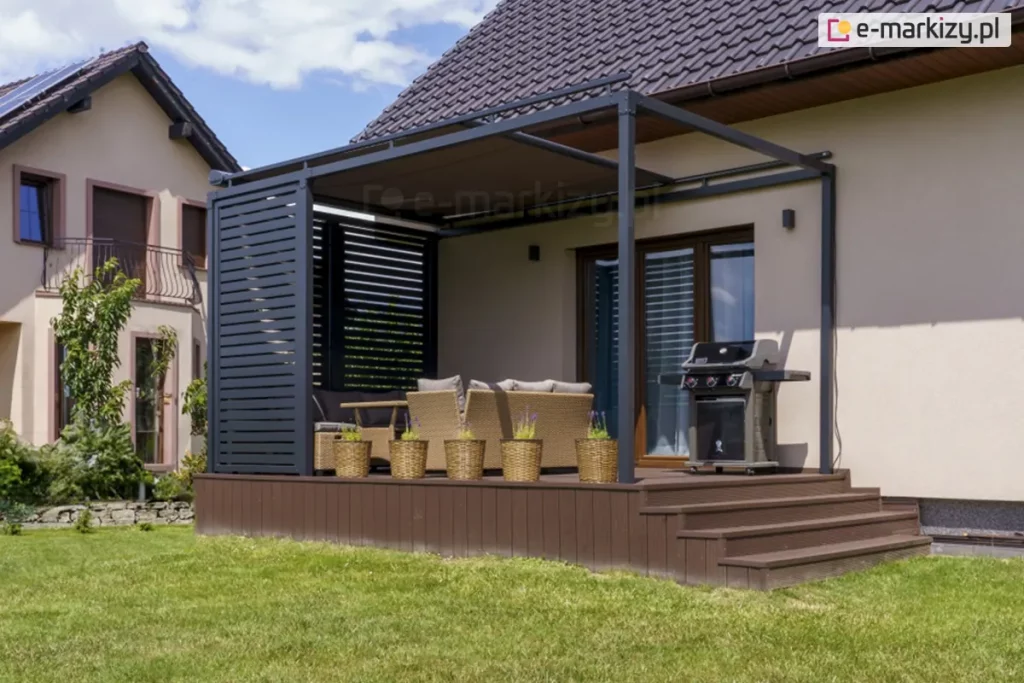 Nowoczesna pergola tarasowa o konstrukcji przyściennej w kolorze antracytowym, zainstalowana na podwyższonym tarasie z brązowych desek kompozytowych. Charakterystycznym elementem jest boczna ściana wykonana z aluminiowych lamel stałych ułożonych poziomo, pełniąca funkcję osłony prywatności i wiatrochronu. Zadaszenie pergoli stanowi refleksol poziomy system veranda z brązową tkaniną akrylową. Na tarasie znajduje się strefa wypoczynkowa z beżowymi meblami rattanowymi oraz profesjonalny grill gazowy ustawiony przy elewacji. Przed narożnikiem pergoli ustawiono pięć wiklinowych donic z roślinami ozdobnymi. Taras posiada szerokie schody prowadzące bezpośrednio na trawnik. W tle widoczny jest sąsiedni dom jednorodzinny z dachem krytym ciemną dachówką oraz zainstalowanymi panelami fotowoltaicznymi. Całość realizacji pokazuje harmonijne połączenie konstrukcji pergoli z bryłą budynku i otaczającym ogrodem.