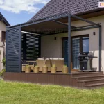 Nowoczesna pergola tarasowa o konstrukcji przyściennej w kolorze antracytowym, zainstalowana na podwyższonym tarasie z brązowych desek kompozytowych. Charakterystycznym elementem jest boczna ściana wykonana z aluminiowych lamel stałych ułożonych poziomo, pełniąca funkcję osłony prywatności i wiatrochronu. Zadaszenie pergoli stanowi refleksol poziomy system veranda z brązową tkaniną akrylową. Na tarasie znajduje się strefa wypoczynkowa z beżowymi meblami rattanowymi oraz profesjonalny grill gazowy ustawiony przy elewacji. Przed narożnikiem pergoli ustawiono pięć wiklinowych donic z roślinami ozdobnymi. Taras posiada szerokie schody prowadzące bezpośrednio na trawnik. W tle widoczny jest sąsiedni dom jednorodzinny z dachem krytym ciemną dachówką oraz zainstalowanymi panelami fotowoltaicznymi. Całość realizacji pokazuje harmonijne połączenie konstrukcji pergoli z bryłą budynku i otaczającym ogrodem.