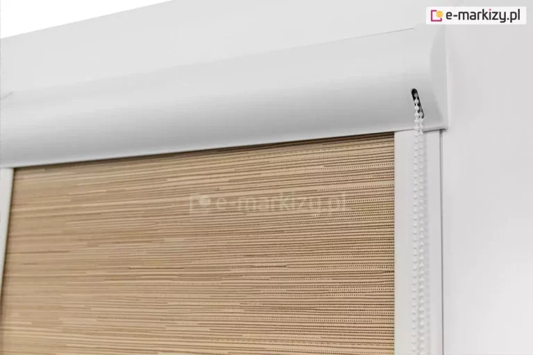 Zbliżenie na górną część systemu roleta lux zamontowanego na białej ramie okiennej. Zdjęcie prezentuje techniczną stronę konstrukcji, skupiając się na białej, aluminiowej kasecie o opływowym kształcie, która maskuje rurę nawojową z tkaniną. Po prawej stronie kasety widoczny jest mechanizm sterujący z precyzyjnym wycięciem, przez które wyprowadzony został biały łańcuszek koralikowy służący do obsługi osłony. Materiał rolety posiada beżową barwę i charakterystyczną, prążkowaną strukturę przypominającą matę bambusową z drobnymi, poziomymi przeplotami ciemniejszej nitki. Tkanina wchodzi bezpośrednio w białe, płaskie prowadnice boczne, które są zamontowane na listwach przy szybowych, co zapewnia idealne napięcie materiału i brak prześwitów przy krawędziach szyby.