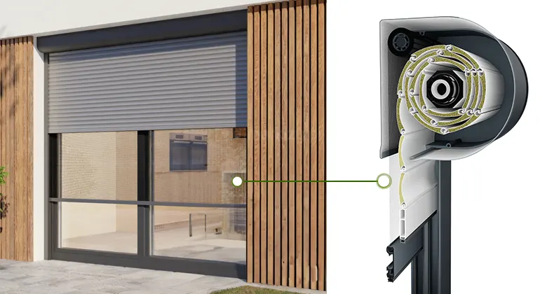 Roleta zewnętrzna z szarym pancerzem i okrągłą skrzynką zamocowana na dużym oknie tarasowym z antracytową ramą, umieszczonym we wnęce okiennej pomiędzy ścianami wyłożonymi drewnianymi lamelami. Roleta wyposażona w opcjonalną zintegrowaną moskitierę, na której umieszczono biały okrągły znacznik, prowadzący prostą zieloną linią na prawo, do znacznika pokazującego moskitierę w przekroju bocznym rolety z okrągłą skrzynką.