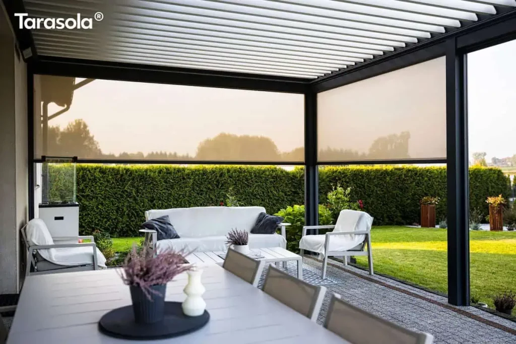 Widok z wnętrza nowoczesnej pergoli tarasowej typu Essential tarasola, zamontowanej przy budynku mieszkalnym. Konstrukcja pergoli wykonana jest z profili aluminiowych w kolorze ciemnego antracytu, a zadaszenie stanowią białe, poziome lamele aluminiowe ustawione pod kątem. Dwa boki pergoli zostały wyposażone w półprzezroczyste rolety zewnętrzne typu screen w kolorze jasnokremowym, które są częściowo opuszczone, przesłaniając widok na ogród i okoliczne drzewa. Wewnątrz zadaszonej przestrzeni znajduje się strefa wypoczynkowa z białą sofą ozdobioną ciemnymi poduszkami oraz dwoma białymi fotelami z narzuconymi jasnymi kocami. Meble ustawione są wokół niskiego, białego stolika kawowego, na którym stoi doniczka z lawendą. Pod meblami wypoczynkowymi leży wzorzysty dywan zewnętrzny w odcieniach błękitu i bieli. Na pierwszym planie widoczny jest fragment białego stołu jadalnianego z dekoracyjną świecą i doniczką z wrzosem na czarnej podkładce. Podłoga tarasu wyłożona jest jasnoszarą kostką brukową. W tle, poza obrębem pergoli, rozciąga się intensywnie zielony, starannie przystrzyżony trawnik oraz wysoki, gęsty żywopłot oddzielający posesję od sąsiedztwa.