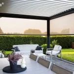 Widok z wnętrza nowoczesnej pergoli tarasowej typu Essential tarasola, zamontowanej przy budynku mieszkalnym. Konstrukcja pergoli wykonana jest z profili aluminiowych w kolorze ciemnego antracytu, a zadaszenie stanowią białe, poziome lamele aluminiowe ustawione pod kątem. Dwa boki pergoli zostały wyposażone w półprzezroczyste rolety zewnętrzne typu screen w kolorze jasnokremowym, które są częściowo opuszczone, przesłaniając widok na ogród i okoliczne drzewa. Wewnątrz zadaszonej przestrzeni znajduje się strefa wypoczynkowa z białą sofą ozdobioną ciemnymi poduszkami oraz dwoma białymi fotelami z narzuconymi jasnymi kocami. Meble ustawione są wokół niskiego, białego stolika kawowego, na którym stoi doniczka z lawendą. Pod meblami wypoczynkowymi leży wzorzysty dywan zewnętrzny w odcieniach błękitu i bieli. Na pierwszym planie widoczny jest fragment białego stołu jadalnianego z dekoracyjną świecą i doniczką z wrzosem na czarnej podkładce. Podłoga tarasu wyłożona jest jasnoszarą kostką brukową. W tle, poza obrębem pergoli, rozciąga się intensywnie zielony, starannie przystrzyżony trawnik oraz wysoki, gęsty żywopłot oddzielający posesję od sąsiedztwa.