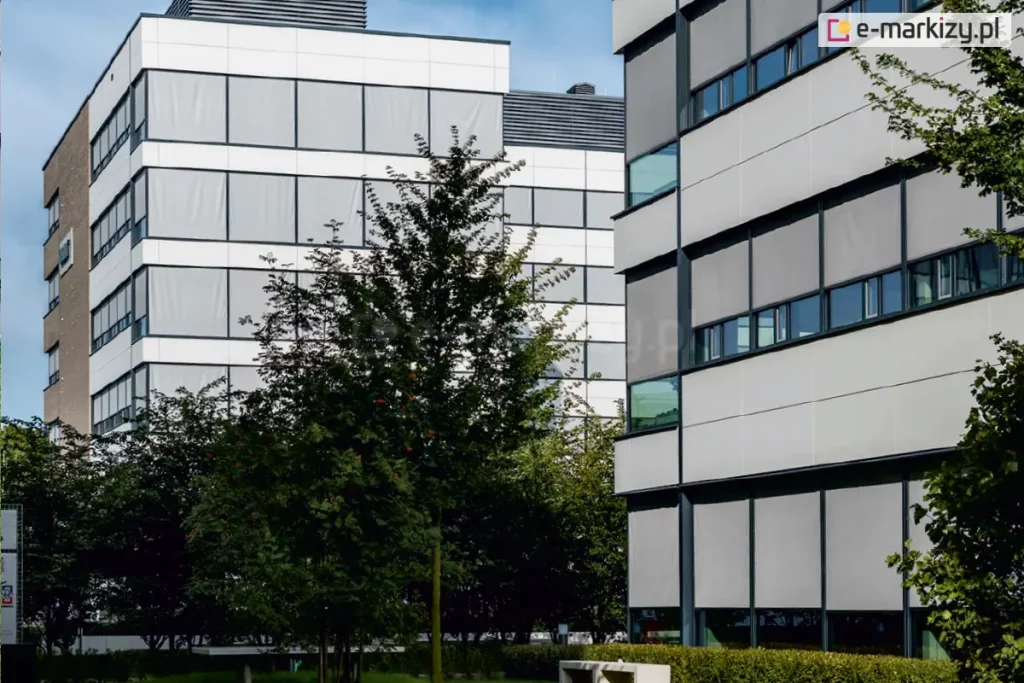 Dwa wysokie biurowce obok siebie, pomiędzy nimi widoczne niskie drzewa oraz zadbany teren zielony otoczony niskim żywopłotem przy którym stoi betonowa ławka. Okna biurowców w większości osłonięte zostały roletami screen o jasnej tkaninie.