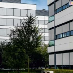 Dwa wysokie biurowce obok siebie, pomiędzy nimi widoczne niskie drzewa oraz zadbany teren zielony otoczony niskim żywopłotem przy którym stoi betonowa ławka. Okna biurowców w większości osłonięte zostały roletami screen o jasnej tkaninie.