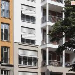 Zdjęcie przedstawia fragment nowoczesnej kamienicy lub budynku wielorodzinnego, na którego elewacji zamontowano kilka systemów osłonowych typu markizoroleta. Budynek posiada białą oraz jasnoszarą fasadę z dużymi przeszkleniami i balkonami. Zastosowane osłony wykonane są z tkaniny w klasyczne, pionowe biało-szare pasy. Trzy widoczne na zdjęciu markizorolety znajdują się w różnym stopniu rozwinięcia: najwyższa i najniższa są opuszczone do połowy okna z wyraźnie odchyloną dolną częścią za pomocą bocznych ramion, natomiast środkowa osłona jest niemal całkowicie zwinięta w kasecie. Konstrukcja ramion pozwala na stworzenie daszka, który chroni wnętrze przed słońcem, jednocześnie umożliwiając naturalną wentylację i widok na zewnątrz. Po prawej stronie kadru widoczne są balkony wsparte na białych kolumnach z ozdobnymi złotymi pierścieniami oraz ciemne, metalowe barierki. W prawej części zdjęcia widoczne są liście i gałęzie drzewa, które rzucają naturalny cień na fragment elewacji.