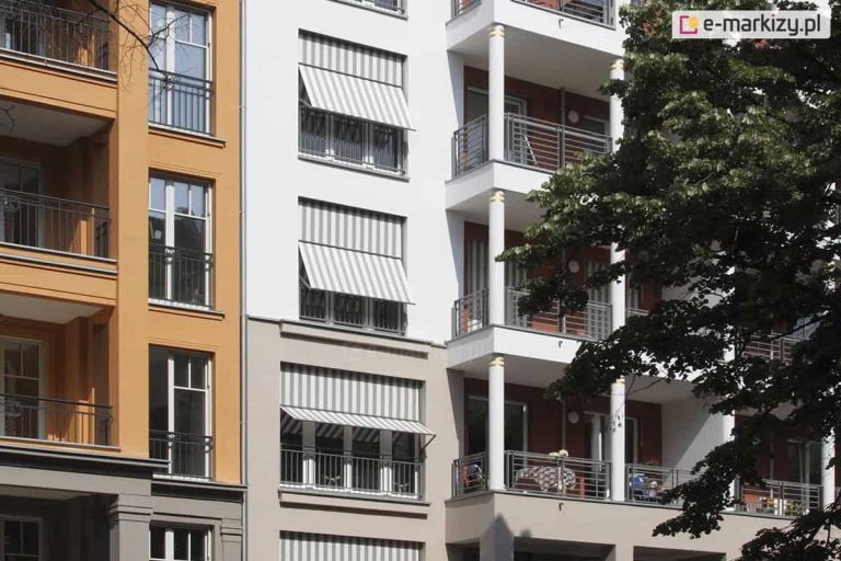Zdjęcie przedstawia fragment nowoczesnej kamienicy lub budynku wielorodzinnego, na którego elewacji zamontowano kilka systemów osłonowych typu markizoroleta. Budynek posiada białą oraz jasnoszarą fasadę z dużymi przeszkleniami i balkonami. Zastosowane osłony wykonane są z tkaniny w klasyczne, pionowe biało-szare pasy. Trzy widoczne na zdjęciu markizorolety znajdują się w różnym stopniu rozwinięcia: najwyższa i najniższa są opuszczone do połowy okna z wyraźnie odchyloną dolną częścią za pomocą bocznych ramion, natomiast środkowa osłona jest niemal całkowicie zwinięta w kasecie. Konstrukcja ramion pozwala na stworzenie daszka, który chroni wnętrze przed słońcem, jednocześnie umożliwiając naturalną wentylację i widok na zewnątrz. Po prawej stronie kadru widoczne są balkony wsparte na białych kolumnach z ozdobnymi złotymi pierścieniami oraz ciemne, metalowe barierki. W prawej części zdjęcia widoczne są liście i gałęzie drzewa, które rzucają naturalny cień na fragment elewacji.