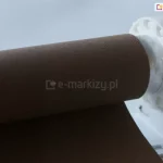Zbliżenie brązowej tkaniny roletowej nawiniętej na rurkę oraz prawego uchwytu rolety PLUS w kolorze białym z widocznym łańcuszkiem do sterowania roletą.