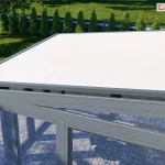widok z góry na nowoczesną konstrukcję ogrodu zimowego lub przeszklonego tarasu, na którego dachu zainstalowano zewnętrzną markizę typu veranda. Poszycie verandy wykonano z tkaniny akrylowej w kolorze białym lub bardzo jasnoszarym jest w pełni rozwinięte, tworząc jednolitą, napiętą płaszczyznę chroniącą wnętrze przed promieniowaniem słonecznym. Tkanina jest osadzona w systemie prowadnic bocznych, a jej krawędź czołowa zakończona jest estetycznym, zaokrąglonym profilem w kolorze antracytowym. Cała konstrukcja nośna obiektu, w tym ramy okienne i profile dachowe, wykonana jest z aluminium lakierowanego na pasujący, ciemnoszary kolor o matowej strukturze. Pod zadaszeniem widoczna jest posadzka wykonana z jasnej kostki brukowej oraz zarys mebli tarasowych. W tle rozpościera się zadbany ogród z zielonym trawnikiem, odgrodzony nowoczesnym płotem z ciemnymi panelami, wzdłuż którego posadzono rząd równo przyciętych tui. Światło dzienne jest rozproszone, co pozwala na wyraźne dostrzeżenie detali technicznych mocowań verandy oraz struktury materiałów użytych do budowy ogrodu zimowego.