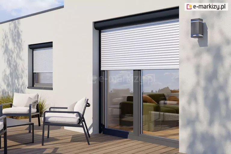 Duże okno tarasowe oraz mniejsze okno pokojowe widoczne w dalszej perspektywie, zamocowane w ścianie o jasnej elewacji, na której po prawej i lewej stronie widoczne są cienie drzew, przesłonięte zostały częściowo opuszczonymi roletami zewnętrznymi w skrzynkach półokrągłych o antracytowym kolorze oraz białych lamelach pancerzy. Na tarasie przed budynkiem, wyłożonym drewnianymi deskami, ustawione zostały trzy krzesła z metalu w kolorze antracytu, wyścielone białymi poduszkami oraz dopasowany do nich niski metalowy stolik.