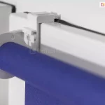 Zbliżenie zamocowania roletki mini w uchwycie bezinwazyjnym zawieszonym na skrzydle okna o białej ramie. Z białymi mocowaniami i mechanizmami roletki kontrastuje chabrowa tkanina.