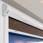 Roleta w kasecie z prowadnicami przestrzennymi Lux Kombi w białym kolorze zamocowana na skrzydle okna o białej ramie, z brązową tkaniną o lekkim wzorze w pionowe, nieregularne linie.