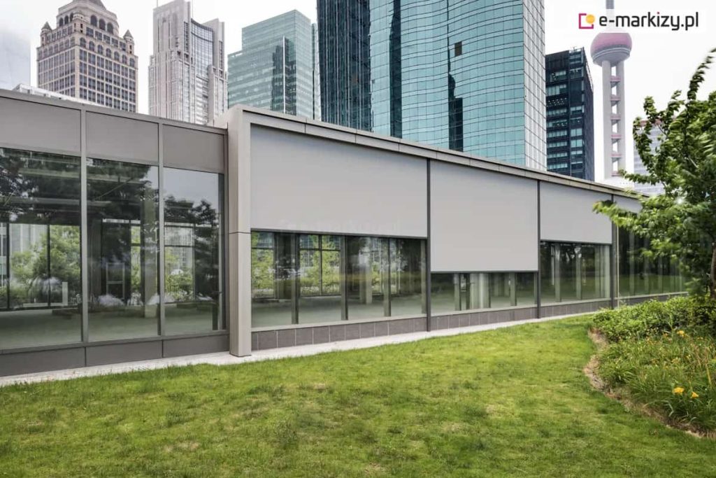 Nowoczesny parterowy budynek biurowy z dużymi przeszklonymi fasadami. Na oknach zamontowane są zewnętrzne rolety screenowe w kolorze jasnoszarym. Rolety te są częściowo rozwinięte, zasłaniając górną część szyb. Ich konstrukcja oparta jest na prostokątnych kasetach zintegrowanych z elewacją, z bocznymi prowadnicami. Tkanina rolet jest lekko przepuszczalna dla światła i odbija refleksy świetlne. W tle widoczne są wysokie, przeszklone wieżowce, co sugeruje miejskie, biznesowe otoczenie. Na pierwszym planie znajduje się zadbany trawnik z niską roślinnością.