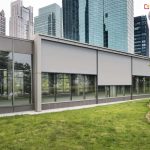 Nowoczesny parterowy budynek biurowy z dużymi przeszklonymi fasadami. Na oknach zamontowane są zewnętrzne rolety screenowe w kolorze jasnoszarym. Rolety te są częściowo rozwinięte, zasłaniając górną część szyb. Ich konstrukcja oparta jest na prostokątnych kasetach zintegrowanych z elewacją, z bocznymi prowadnicami. Tkanina rolet jest lekko przepuszczalna dla światła i odbija refleksy świetlne. W tle widoczne są wysokie, przeszklone wieżowce, co sugeruje miejskie, biznesowe otoczenie. Na pierwszym planie znajduje się zadbany trawnik z niską roślinnością.