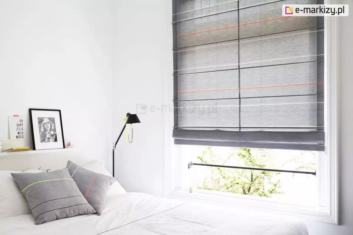 Jasna i minimalistyczna sypialnia, w której głównym elementem dekoracyjnym okna są nowoczesne rolety rzymskie. Tkanina osłony ma kolor grafitowy o melanżowym splocie, a jej unikalny design podkreślają cienkie, poziome pasy w kolorach czerwonym, żółtym i białym, które przebiegają w regularnych odstępach na wysokości fiszbin. Rolety rzymskie są częściowo podciągnięte, tworząc u dołu grubą, estetyczną zakładkę z materiału, co odsłania dolną część okna oraz widok na zielone korony drzew na zewnątrz. Sypialnia urządzona jest w bieli, co potęguje wrażenie czystości i doświetlenia. Po lewej stronie znajduje się łóżko z białą pościelą, na którym ułożono dwie szare poduszki dekoracyjne. Wzór na poduszkach – poziome linie w kolorach neonowej żółci i czerwieni – idealnie koresponduje z detalami widocznymi na materiale, z którego wykonano rolety rzymskie. Nad wezgłowiem łóżka zamontowano wąską, białą półkę, na której stoi czarno-biała fotografia w ramce oraz grafika z napisem. Obok łóżka stoi smukła, czarna lampka nocna o regulowanym ramieniu, ozdobiona jaskrawożółtym elementem. Całość aranżacji stanowi spójną kompozycję, w której surowość szarości i bieli została przełamana energetycznymi akcentami kolorystycznymi obecnymi na tekstyliach okiennych.