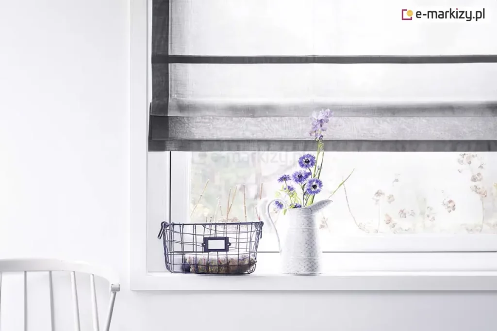 Jasne i minimalistyczne wnętrze, w którym głównym punktem kompozycji jest okno ozdobione przez eleganckie rolety rzymskie. Tkanina osłony jest biała i transparentna, o delikatnej strukturze przypominającej firankę, co pozwala na maksymalne doświetlenie pomieszczenia przy jednoczesnym zachowaniu prywatności. Wyjątkowy charakter tej rolety rzymskiej podkreślają szerokie, poziome pasy oraz pionowe obramowanie wykonane z materiału w kolorze ciemnej, nasyconej szarości lub grafitu. Mechanizm jest częściowo podciągnięty, przez co u dołu tworzy się estetyczna, warstwowa zakładka z materiału. Okno osadzone jest w prostej, białej ramie, która zlewa się z gładką powierzchnią białej ściany. Na szerokim, białym parapecie ustawiono dekoracyjne przedmioty: biały, ceramiczny dzbanek pełniący funkcję wazonu z bukietem niebieskich i fioletowych kwiatów polnych oraz metalowy, ażurowy koszyk w kolorze czarnym, wewnątrz którego znajduje się paczka zawinięta w ozdobny papier. Po lewej stronie, w dolnym rogu zdjęcia, widoczny jest fragment białego, klasycznego krzesła szczebelkowego. Przez transparentny materiał, z którego wykonano rolety rzymskie, prześwituje rozmyty widok ogrodu, tworząc spokojną i świeżą atmosferę domowego wnętrza.