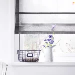 Jasne i minimalistyczne wnętrze, w którym głównym punktem kompozycji jest okno ozdobione przez eleganckie rolety rzymskie. Tkanina osłony jest biała i transparentna, o delikatnej strukturze przypominającej firankę, co pozwala na maksymalne doświetlenie pomieszczenia przy jednoczesnym zachowaniu prywatności. Wyjątkowy charakter tej rolety rzymskiej podkreślają szerokie, poziome pasy oraz pionowe obramowanie wykonane z materiału w kolorze ciemnej, nasyconej szarości lub grafitu. Mechanizm jest częściowo podciągnięty, przez co u dołu tworzy się estetyczna, warstwowa zakładka z materiału. Okno osadzone jest w prostej, białej ramie, która zlewa się z gładką powierzchnią białej ściany. Na szerokim, białym parapecie ustawiono dekoracyjne przedmioty: biały, ceramiczny dzbanek pełniący funkcję wazonu z bukietem niebieskich i fioletowych kwiatów polnych oraz metalowy, ażurowy koszyk w kolorze czarnym, wewnątrz którego znajduje się paczka zawinięta w ozdobny papier. Po lewej stronie, w dolnym rogu zdjęcia, widoczny jest fragment białego, klasycznego krzesła szczebelkowego. Przez transparentny materiał, z którego wykonano rolety rzymskie, prześwituje rozmyty widok ogrodu, tworząc spokojną i świeżą atmosferę domowego wnętrza.