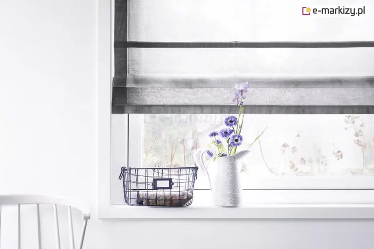 Jasne i minimalistyczne wnętrze, w którym głównym punktem kompozycji jest okno ozdobione przez eleganckie rolety rzymskie. Tkanina osłony jest biała i transparentna, o delikatnej strukturze przypominającej firankę, co pozwala na maksymalne doświetlenie pomieszczenia przy jednoczesnym zachowaniu prywatności. Wyjątkowy charakter tej rolety rzymskiej podkreślają szerokie, poziome pasy oraz pionowe obramowanie wykonane z materiału w kolorze ciemnej, nasyconej szarości lub grafitu. Mechanizm jest częściowo podciągnięty, przez co u dołu tworzy się estetyczna, warstwowa zakładka z materiału. Okno osadzone jest w prostej, białej ramie, która zlewa się z gładką powierzchnią białej ściany. Na szerokim, białym parapecie ustawiono dekoracyjne przedmioty: biały, ceramiczny dzbanek pełniący funkcję wazonu z bukietem niebieskich i fioletowych kwiatów polnych oraz metalowy, ażurowy koszyk w kolorze czarnym, wewnątrz którego znajduje się paczka zawinięta w ozdobny papier. Po lewej stronie, w dolnym rogu zdjęcia, widoczny jest fragment białego, klasycznego krzesła szczebelkowego. Przez transparentny materiał, z którego wykonano rolety rzymskie, prześwituje rozmyty widok ogrodu, tworząc spokojną i świeżą atmosferę domowego wnętrza.