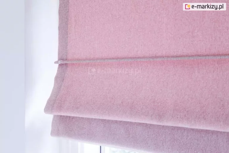 Zbliżenie na fragment podciągniętej rolety rzymskiej wykonanej z miękkiej, teksturowanej tkaniny w kolorze jasnego, cukierkowego różu. Materiał charakteryzuje się gęstym splotem o delikatnie melanżowej strukturze, która nadaje mu wizualną głębię i matowe wykończenie. Na zdjęciu widoczny jest boczny brzeg rolety z estetycznym obszyciem oraz pozioma fiszbina umieszczona w specjalnym tunelu, która odpowiada za precyzyjne i równe składanie się materiału. Dolna część tkaniny tworzy miękką, podwójną zakładkę, typową dla mechanizmu rzymskiego, co nadaje dekoracji okiennej trójwymiarowości. Oświetlenie jest jasne i naturalne, podkreślając puszystość włókien oraz subtelne cienie powstające na zgięciach materiału. Tło po lewej stronie stanowi gładka, biała ściana, która kontrastuje z ciepłym odcieniem tkaniny, natomiast u dołu widać fragment ramy okiennej, sugerujący montaż rolety w świetle okna.