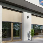 Fragment parterowej części elewacji budynku w stylu modernistycznym z dużymi przeszkleniami tarasowymi, zasłoniętymi zewnętrznymi osłonami Refleksol 90 Selt. System screenowy zastosowany na zdjęciu wyposażony jest w prowadnice boczne i estetyczne, zaokrąglone kasety, które harmonijnie łączą się z bryłą budynku. Jasnobeżowa tkanina screenowa, o drobno tkanej strukturze, częściowo zacienia okna, tworząc delikatny cień wewnątrz pomieszczeń. Dolna część szyb pozostaje odkryta, umożliwiając widok na zewnątrz – widać przez nie nowoczesne, drewniane meble oraz zieloną roślinność w ogrodzie. Elewacja utrzymana w jasnych kolorach kontrastuje z ciemnymi akcentami kaset i obramowań okiennych, a całość kompozycji podkreśla funkcjonalny i jednocześnie estetyczny charakter zastosowanych rozwiązań przeciwsłonecznych. Rośliny doniczkowe i cień rzucony przez liście na elewację dodają naturalnego charakteru przestrzeni tarasowej.