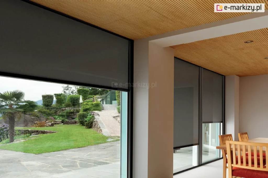 Jasne, eleganckie wnętrze jadalni z drewnianym sufitem wykończonym lamelami oraz meblami w naturalnym odcieniu, skontrastowane z ciemnoszarymi roletami screenowymi na dużych przeszkleniach. Ciemna tkanina Refleksoli dodaje głębi, jednocześnie zachowując lekkość i nowoczesność przestrzeni. Przez przeszklone ściany widoczny jest starannie zaprojektowany ogród z przystrzyżonymi krzewami, kamiennymi murkami i egzotyczną roślinnością. Minimalistyczne, cienkie ramy okienne harmonizują z roletami, a całość emanuje spokojem, elegancją i funkcjonalnym podejściem do projektowania wnętrz.