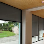 Jasne, eleganckie wnętrze jadalni z drewnianym sufitem wykończonym lamelami oraz meblami w naturalnym odcieniu, skontrastowane z ciemnoszarymi roletami screenowymi na dużych przeszkleniach. Ciemna tkanina Refleksoli dodaje głębi, jednocześnie zachowując lekkość i nowoczesność przestrzeni. Przez przeszklone ściany widoczny jest starannie zaprojektowany ogród z przystrzyżonymi krzewami, kamiennymi murkami i egzotyczną roślinnością. Minimalistyczne, cienkie ramy okienne harmonizują z roletami, a całość emanuje spokojem, elegancją i funkcjonalnym podejściem do projektowania wnętrz.