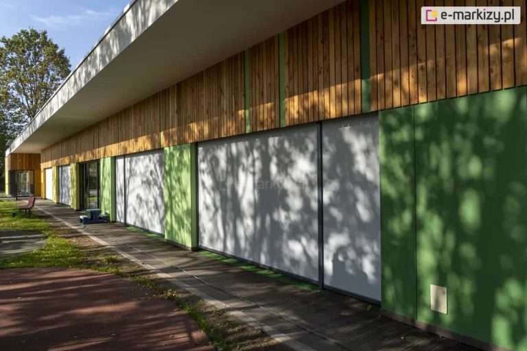 Nowoczesny budynek parterowy z dużymi przeszkleniami, osłoniętymi zewnętrznymi roletami screenowymi w kolorze jasnoszarym. Rolety są całkowicie opuszczone i zamontowane w prowadnicach bocznych, zapewniających stabilność materiału. Fasada budynku wykończona jest zielonymi panelami i drewnianą okładziną pionową w górnej części. Taras wyłożony jest płytami betonowymi, a w otoczeniu widoczne są elementy placu zabaw oraz ławki. Całość wskazuje na obiekt użyteczności publicznej, np. przedszkole lub centrum edukacyjne.