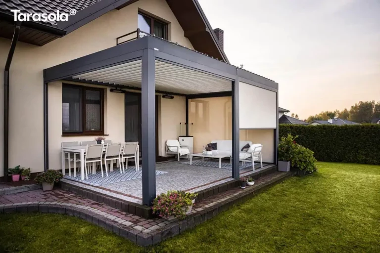 Nowoczesna zadaszona konstrukcja tarasowa typu pergola w kolorze ciemnoszarym antacytowym przylegająca bezpośrednio do ściany piętrowego domu jednorodzinnego o jasnej elewacji. Aluminiowa konstrukcja wspiera dach wykonany z ruchomych białych lameli, które są częściowo otwarte. Pod zadaszeniem znajduje się przestronna strefa wypoczynkowa i jadalniana urządzona na podwyższonym tarasie wyłożonym jasną kostką brukową i otoczonym ciemniejszą krawędzią z kostki. Po lewej stronie pod oknem domu stoi biały prostokątny stół z sześcioma dopasowanymi białymi krzesłami ustawionymi na niebiesko-białym wzorzystym dywanie zewnętrznym. Po prawej stronie w głębi znajduje się strefa relaksu z białą sofą, fotelem oraz niskim stolikiem kawowym, również umieszczona na dekoracyjnym dywanie. Jeden z bocznych segmentów pergoli został wyposażony w pionową białą roletę typu screen, która jest częściowo opuszczona, zapewniając ochronę przed słońcem i wiatrem. Na elewacji budynku pod dachem pergoli zamontowano promienniki ciepła oraz oświetlenie punktowe. Całość otacza zadbany zielony trawnik, a na obrzeżach tarasu oraz wzdłuż ścieżki z kostki brukowej ustawiono liczne donice z kwitnącymi roślinami w odcieniach różu i fioletu. W tle widoczny jest wysoki żywopłot oraz fragmenty sąsiedniej zabudowy pod jasnym, popołudniowym niebem.