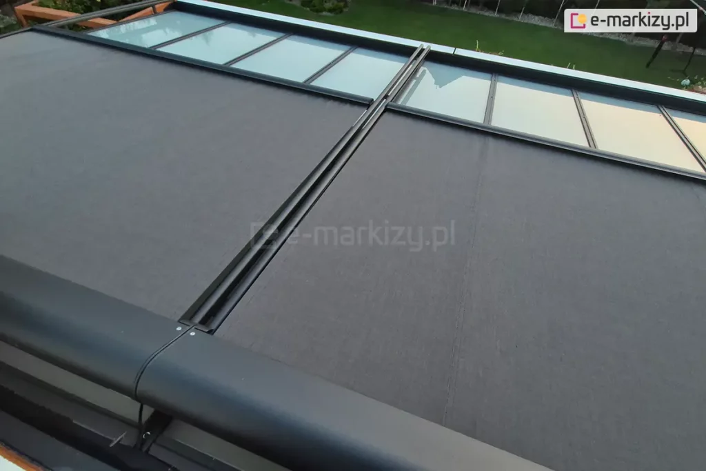 Zdjęcie przedstawia widok z góry na system dwumodułowej markizy werandowa, zamontowanej na aluminiowej konstrukcji ogrodu zimowego. Poszycie markizy wykonane jest z wysokiej jakości tkaniny akrylowej o gęstym splocie w kolorze ciemnoszarym (antracytowym). System jest w fazie częściowego rozwinięcia – dwa segmenty materiału są wysunięte do połowy, odsłaniając w górnej części kadru przeszklone panele dachu ujęte w czarne ramy. Elementy konstrukcyjne, w tym zaokrąglona kaseta osłonowa, prowadnice boczne oraz belki frontowe, są lakierowane proszkowo na kolor matowego antracytu. W tle widoczny jest fragment zadbanego ogrodu z zielonym trawnikiem, drzewami liściastymi oraz drewnianą konstrukcją pergoli po lewej stronie. Perspektywa zdjęcia z góry pozwala na dokładne przyjrzenie się mechanizmowi prowadzenia tkaniny oraz estetyce wykończenia detali.
