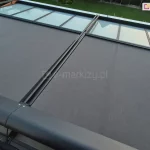 Zdjęcie przedstawia widok z góry na system dwumodułowej markizy werandowa, zamontowanej na aluminiowej konstrukcji ogrodu zimowego. Poszycie markizy wykonane jest z wysokiej jakości tkaniny akrylowej o gęstym splocie w kolorze ciemnoszarym (antracytowym). System jest w fazie częściowego rozwinięcia – dwa segmenty materiału są wysunięte do połowy, odsłaniając w górnej części kadru przeszklone panele dachu ujęte w czarne ramy. Elementy konstrukcyjne, w tym zaokrąglona kaseta osłonowa, prowadnice boczne oraz belki frontowe, są lakierowane proszkowo na kolor matowego antracytu. W tle widoczny jest fragment zadbanego ogrodu z zielonym trawnikiem, drzewami liściastymi oraz drewnianą konstrukcją pergoli po lewej stronie. Perspektywa zdjęcia z góry pozwala na dokładne przyjrzenie się mechanizmowi prowadzenia tkaniny oraz estetyce wykończenia detali.